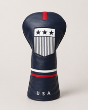 Navy USA-inspired driver headcover photographed upright on a clean warm gray studio background. The leather-style cover features a red top tab, embroidered white crest with three stars and vertical stripe detailing, white and red bands around the middle, and “USA” lettering near the bottom.