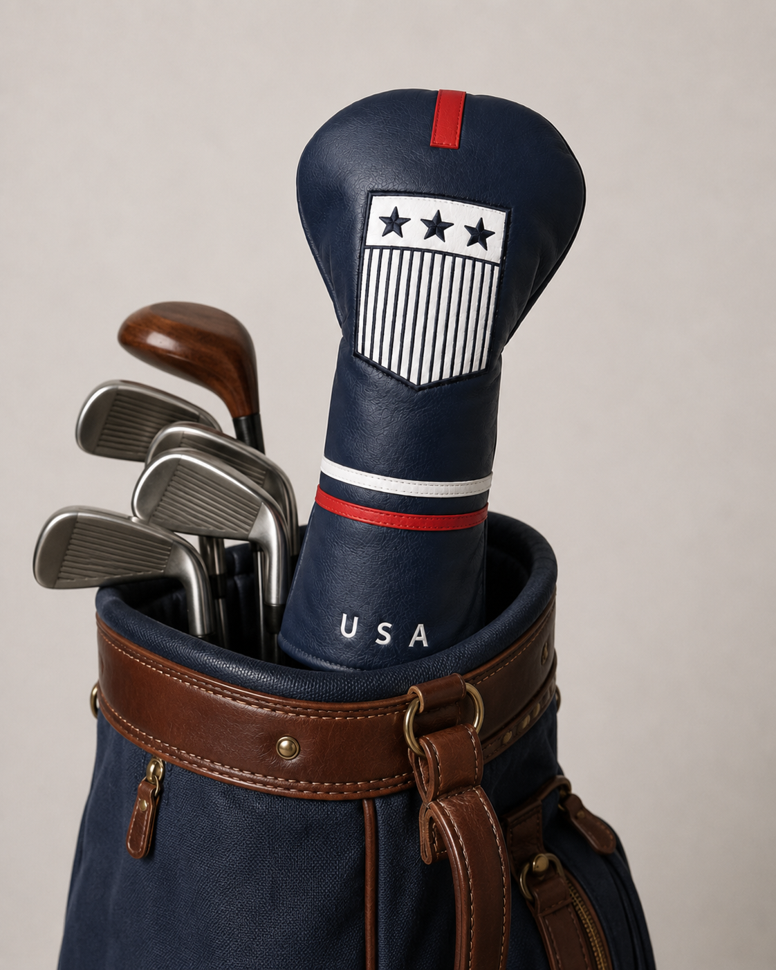 USA driver headcover displayed on a club inside a vintage navy and brown golf bag. The navy leather-style cover features a red top tab, embroidered white crest with stars and stripes, red and white banding, and “USA” lettering. Classic irons and wood-style clubs sit behind it.