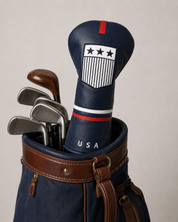 USA driver headcover displayed on a club inside a vintage navy and brown golf bag. The navy leather-style cover features a red top tab, embroidered white crest with stars and stripes, red and white banding, and “USA” lettering. Classic irons and wood-style clubs sit behind it.